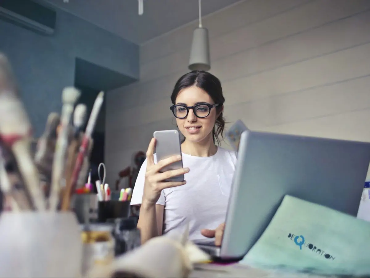 Mulher de óculos lendo uma mensagem no celular, em frente ao laptop
