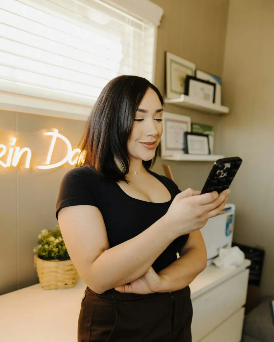 Mulher sorrindo enquanto olha o celular, em pé num escritório iluminado