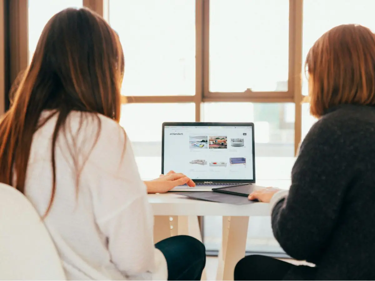 Duas mulheres conversando em frente a um laptop que mostra um site