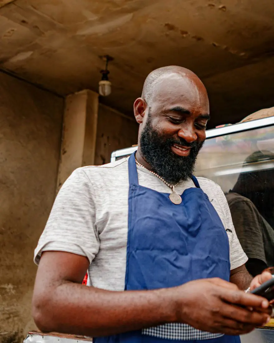 Homem de avental sorrindo enquanto olha o celular, atrás do balcão de um comércio de alimentos